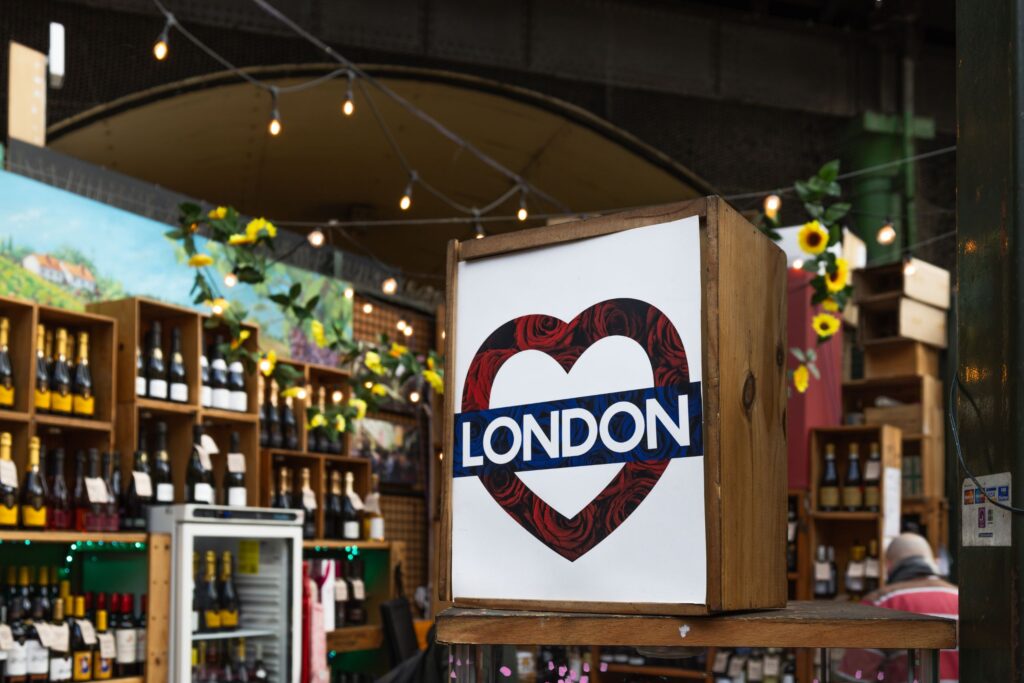 Wood-framed sign with a London heart graphic at a market stall, shelves of bottles behind and string lights overhead