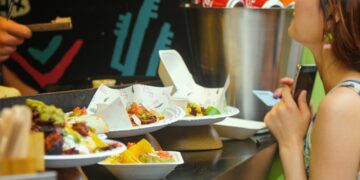woman-eating-street-food-london-market-unsplash - LONDONwebzine A woman sampling street food at a busy London neighbourhoods market, surrounded by global food stalls
