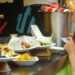 woman-eating-street-food-london-market-unsplash - LONDONwebzine A woman sampling street food at a busy London neighbourhoods market, surrounded by global food stalls