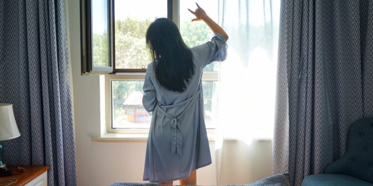 A woman standing by a bright London window in the morning, building a healthy sleep routine through early light exposure