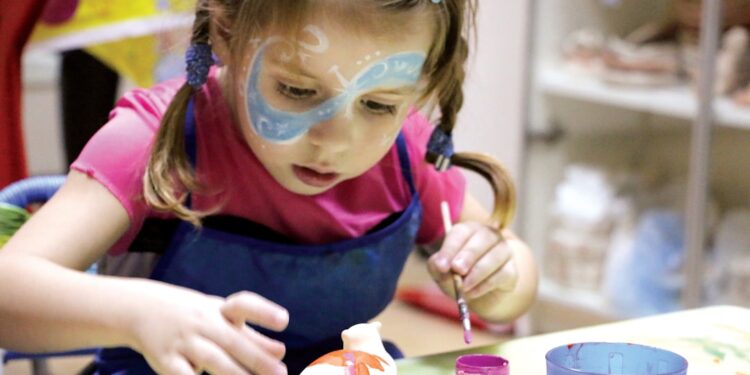 child-painting-pottery-class-unsplash - LONDONwebzine A young child concentrating on a pottery wheel during kids workshops in London, guided by an art instructor in a studio.